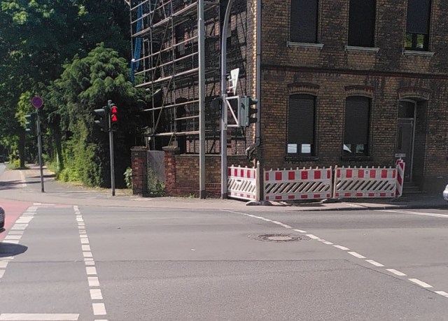 An einer Ampelkreuzung ist der schmale Gehweg vollständig gesperrt durch eine Baustelle. Eine Beschilderung gibt es nicht