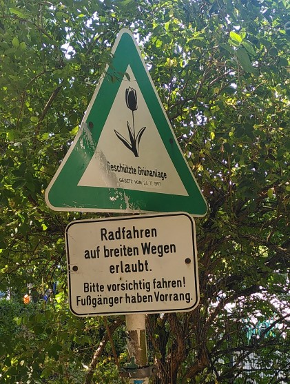 Zwei Schilder, oben grünes Dreieck mit Spitze nach oben, stilisierte Tulpe und dem Schriftzug "Geschützte Grünanlage" und darunter ein Hinweisschild: "Radfahren auf breiten Wegen erlaubt. Bitte vorsichtig fahren! Fußgänger haben Vorrang."
