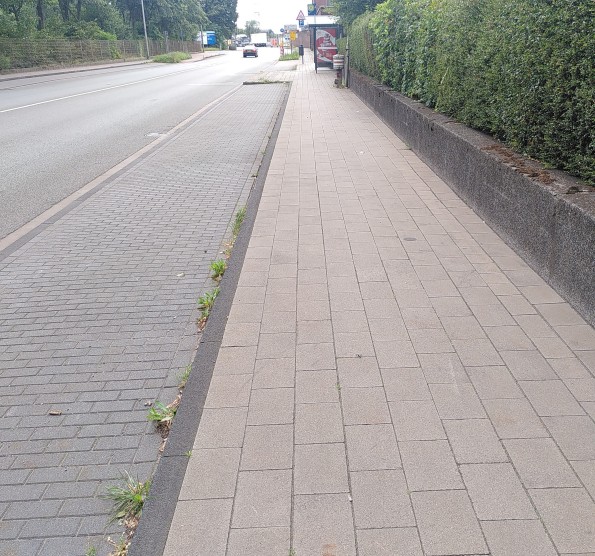 Links ein leerer Parkstreifen, rechts ein schmaler Gehweg, im Hintergrund eine Bushaltestelle.