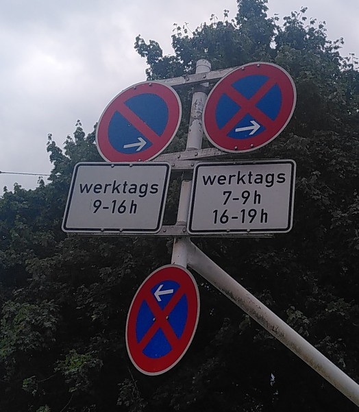 Zu sehen sind drei Schilder, zwei davon mit Zusatzzeichen an einem langen Mast. Oben Halteverbot und absolutes Halteverbot zu bestimmten Zeiten nach Rechts. Unten absolutes Halteverbot nach Links.