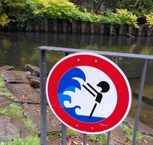 Vor der Wupper steht ein Zaun mit einem runden Schild mit rotem Rand, stilisierter Flutwelle und einer Person.