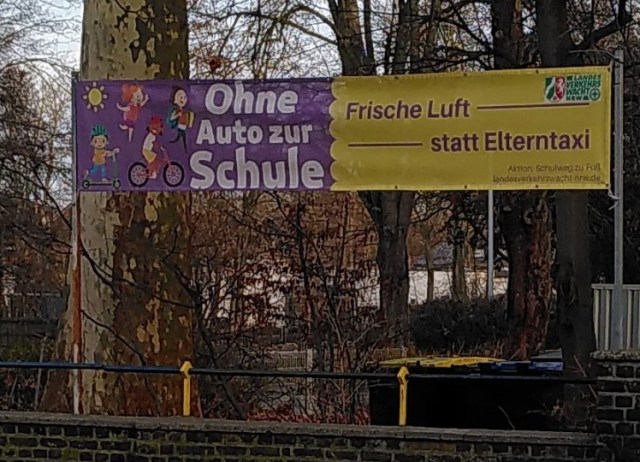 Schulgebäude mit Banner der Landesverkehrswacht NRW davor, darauf steht: "Ohne Auto zur Schule. Frische Luft statt Elterntaxi.  Aktion: Schulweg zu Fuß"