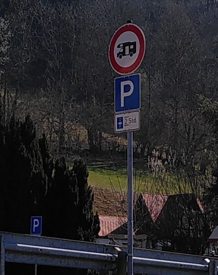 Im Hintergrund bewaldeter Hand, Vordergrund ein Schild mit Durchfahrtsverbot für Wohnmobil darunter Parkplatz und Parkscheibe