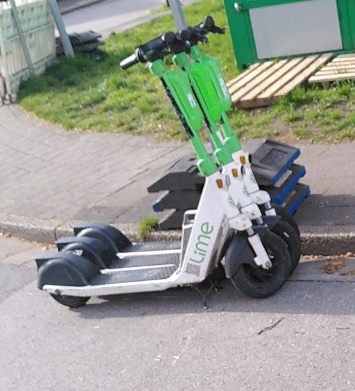 Drei Ihscooter der Firma LIME parken nebeneinander entgegen der Fahrtrichtung kurz vor einer Einmündung.