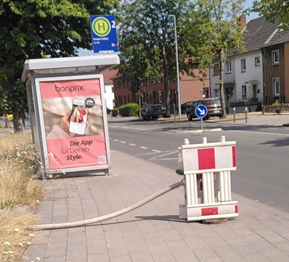 Von Links nach Rechts. Grundstück, ein dicker Wasserschlauch liegt über den Gehweg, angeschlossen an einen Hydranten, der fast am Fahrbahnrand steht. Im Hintergrund eine Bushaltestelle.